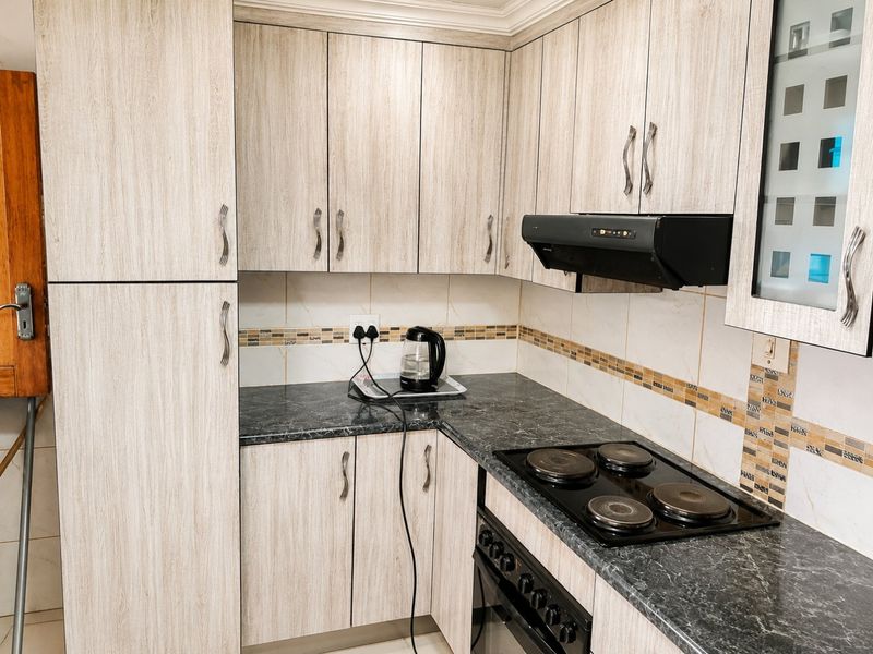 A compact kitchen featuring light wood-grain cabinetry, dark laminate countertops, a built-in oven, electric hob, and a tiled backsplash.