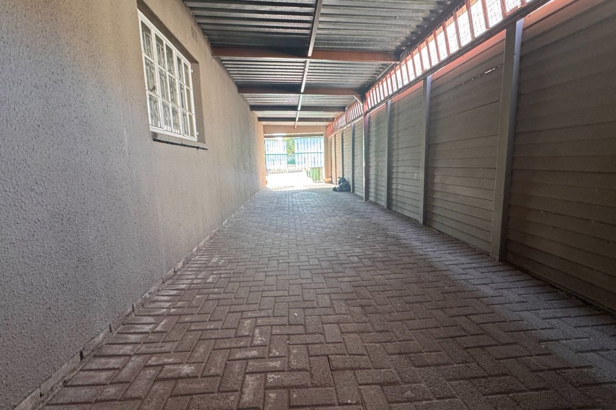 A covered side walkway or driveway area featuring paved flooring, a corrugated metal roof structure, and a secure boundary wall.