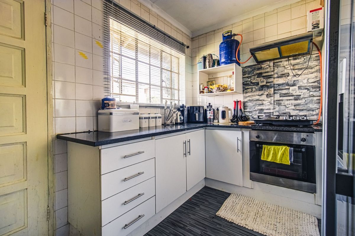 A compact kitchen featuring white cabinetry, granite countertops, a built-in oven with gas hob, and tiled splashbacks, requiring some cosmetic updates.
