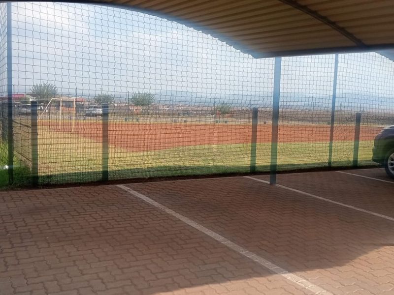 A covered parking bay featuring paved flooring and security mesh fencing, overlooking an adjacent sports field.