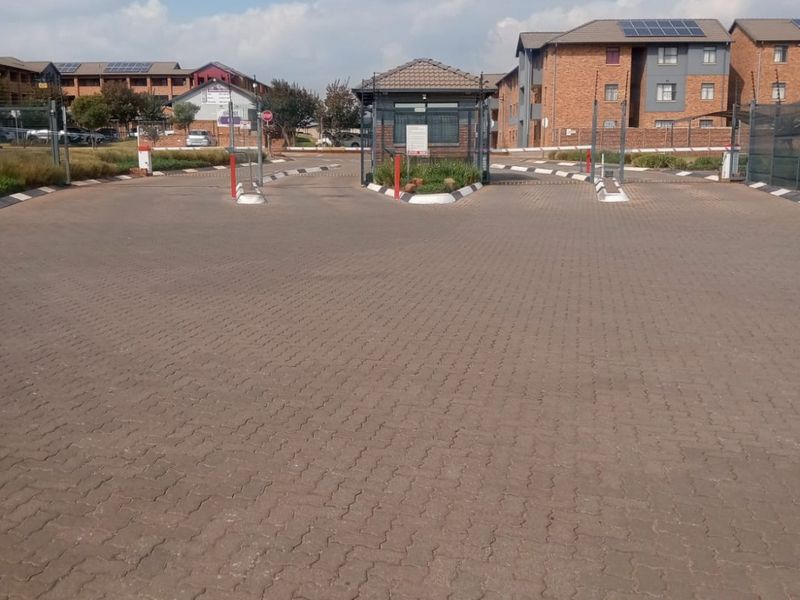 The main entrance to the apartment complex featuring a security guard house, paved driveway, and controlled access boom gates.