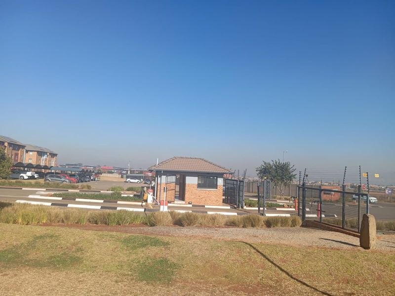 View of the apartment complex entrance featuring a security guard house, access control gates, and paved parking areas with carports.