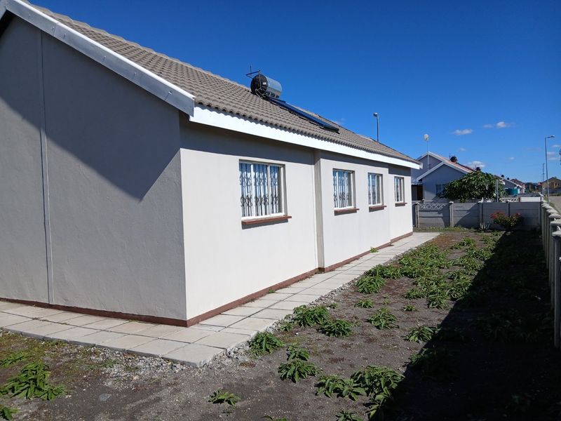 Rear view of the house featuring a solar water heater, tiled roof, security bars on windows, and a paved walkway alongside an unlandscaped garden area.