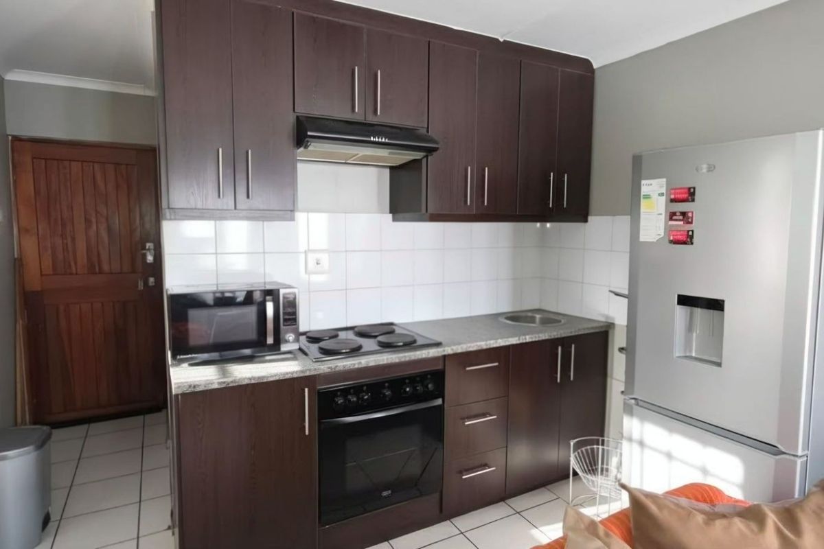 A compact kitchen featuring dark wood cabinetry, tiled splashbacks, a built-in oven, hob, and extractor fan, with tiled flooring throughout.