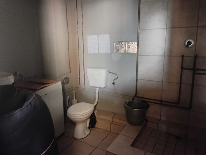 A dated bathroom featuring exposed plumbing, tiled surfaces, and a basic toilet. The space requires renovation to improve functionality and aesthetic appeal.
