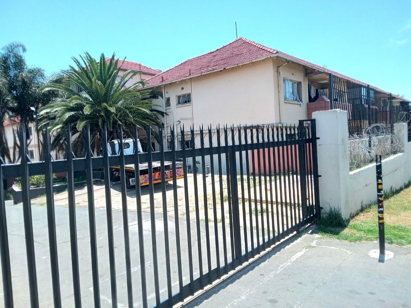Exterior view of an apartment building featuring a metal security gate, perimeter wall with razor wire, and a tiled roof structure.