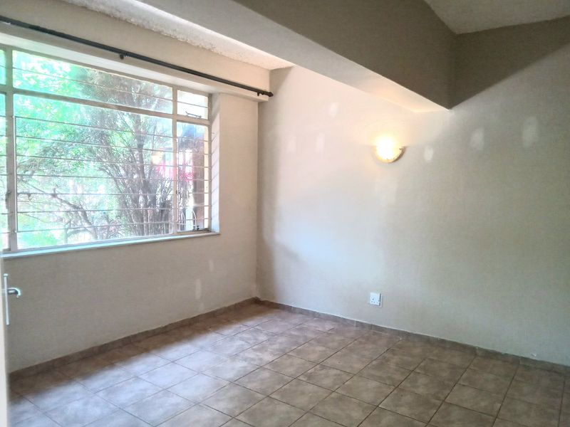 An empty living room featuring tiled flooring, a large window with security bars, and wall-mounted lighting, currently requiring cosmetic wall repairs and fresh paint.