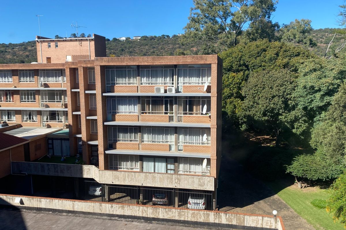 Exterior view of a multi-story brick apartment building featuring balconies, sliding windows, and a ground-level covered parking area set against a wooded backdrop.