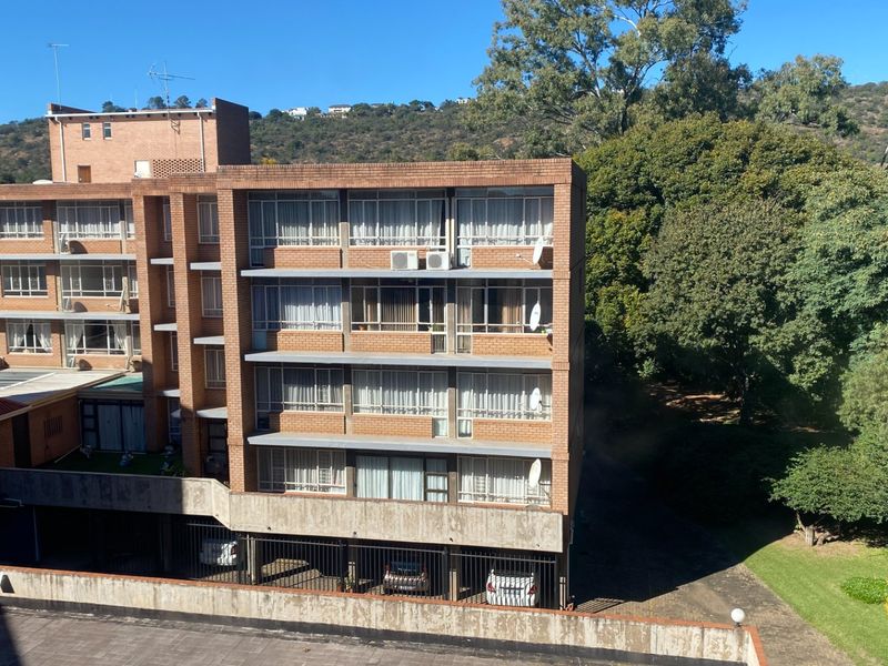Exterior view of a multi-story brick apartment building featuring balconies, sliding windows, and a ground-level covered parking area set against a wooded backdrop.