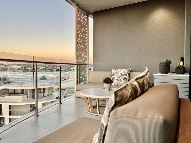 A modern covered balcony featuring a glass balustrade, decorative privacy screen, and tiled flooring, offering expansive urban views.