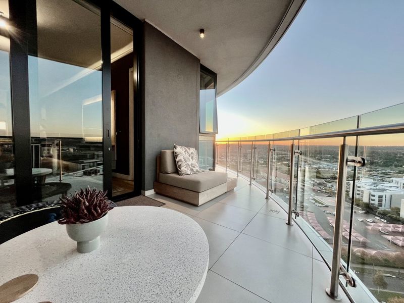A modern, spacious balcony featuring a sleek glass balustrade, tiled flooring, and expansive sliding glass doors offering panoramic sunset views.