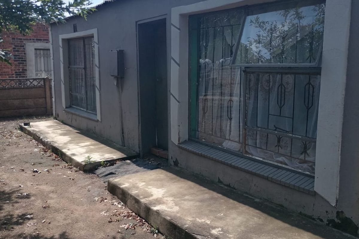 Rear exterior view of a single-story house featuring plastered walls, metal security bars on windows, and a concrete walkway.