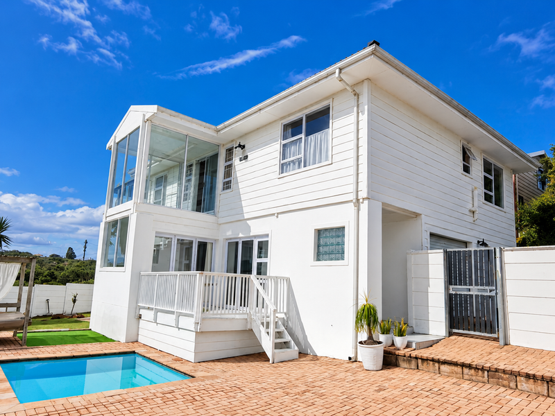 Rear view of a two-story house featuring a swimming pool, raised timber deck, paved patio area, and white weatherboard exterior cladding.