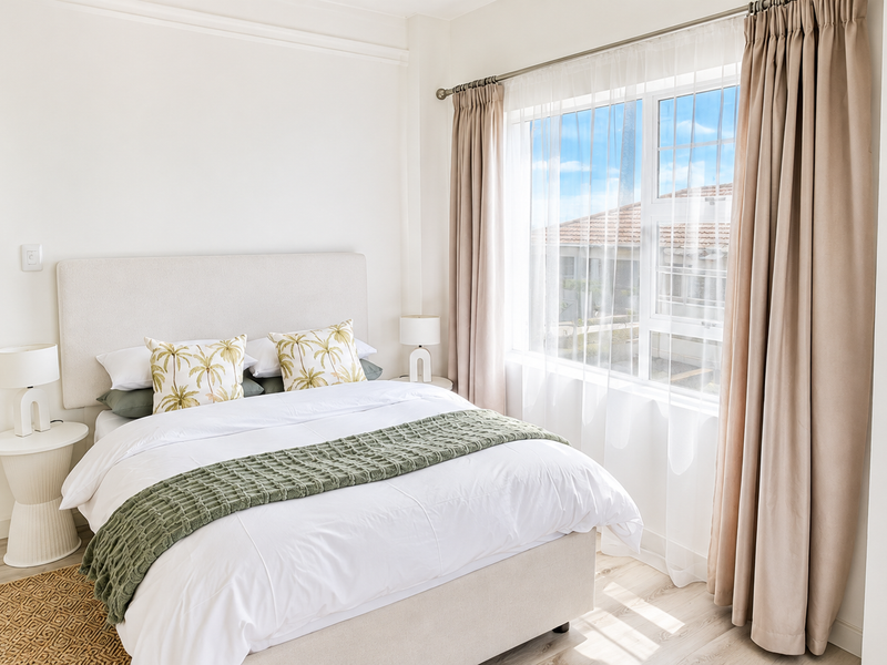 A bright and well-maintained bedroom featuring light-toned laminate flooring and a large window with neutral curtains.