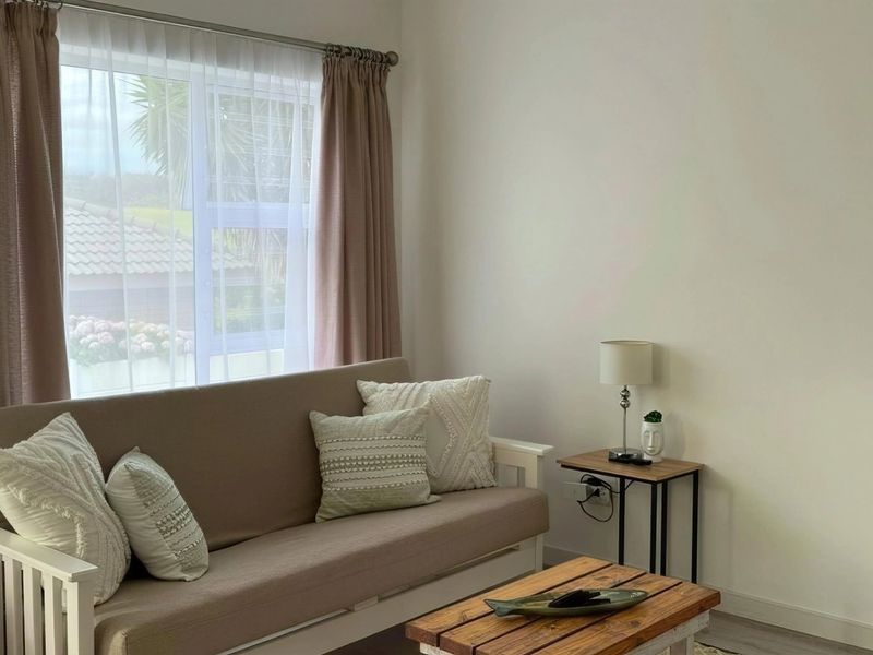A bright living room featuring neutral walls, a large window with curtains, and a doorway leading to an adjacent bedroom.
