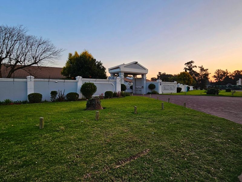 A well-maintained gated entrance to the townhouse complex featuring a paved driveway, perimeter wall with electric fencing, and manicured lawn.