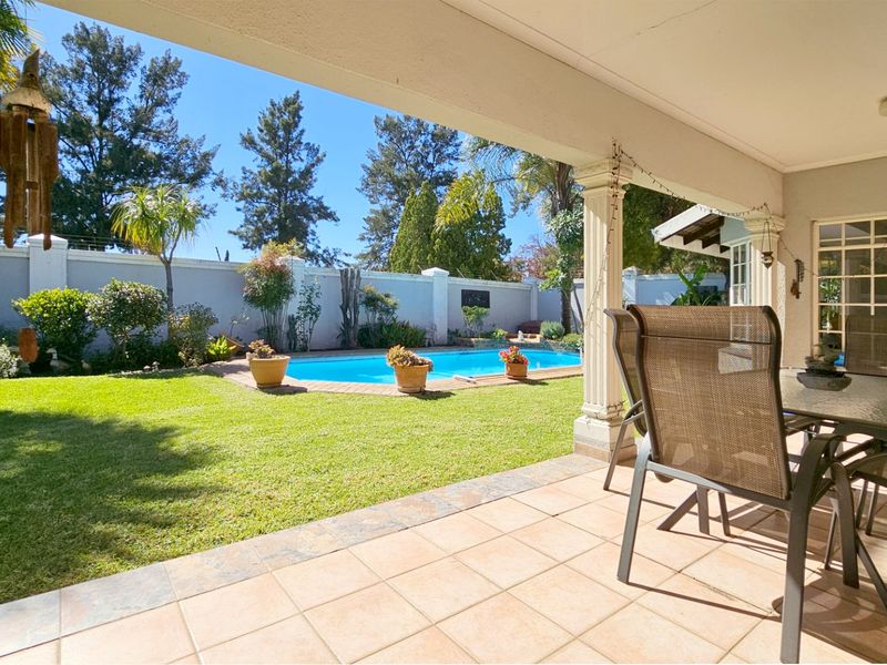 A covered patio overlooking a private garden and swimming pool, featuring tiled flooring and structural columns.