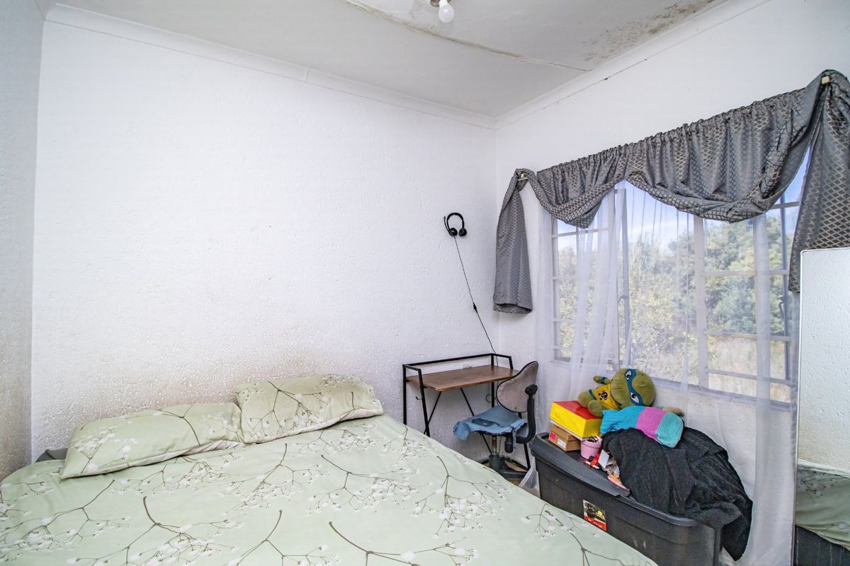 A bedroom featuring a window with curtains and visible signs of ceiling and wall surface deterioration requiring maintenance.