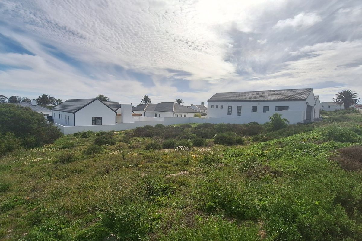 A view of a vacant residential plot featuring surrounding modern white homes with tiled roofs and boundary walls in a quiet neighborhood.