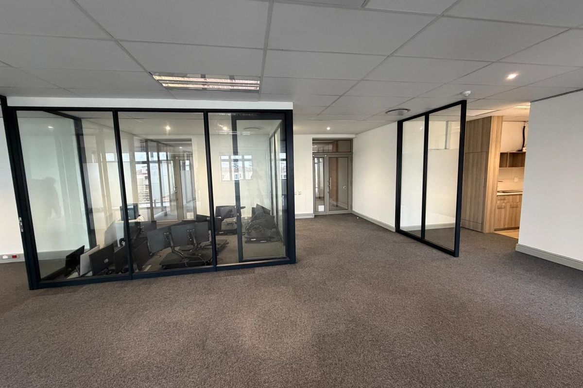 Modern commercial office space featuring glass-partitioned meeting rooms, carpeted flooring, suspended ceiling tiles, and an integrated kitchenette area.