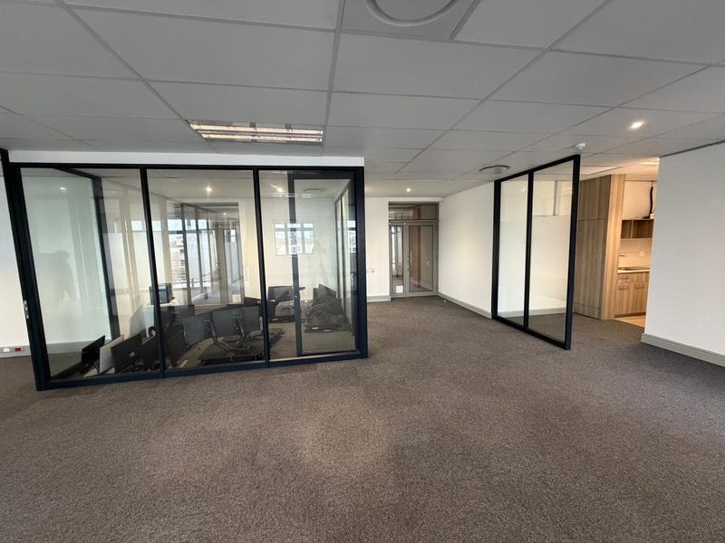 Modern commercial office space featuring glass-partitioned meeting rooms, carpeted flooring, suspended ceiling tiles, and an integrated kitchenette area.