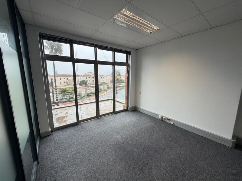 A bright, empty commercial office space featuring grey carpeting, a suspended ceiling with recessed lighting, and large glass doors leading to a private balcony.