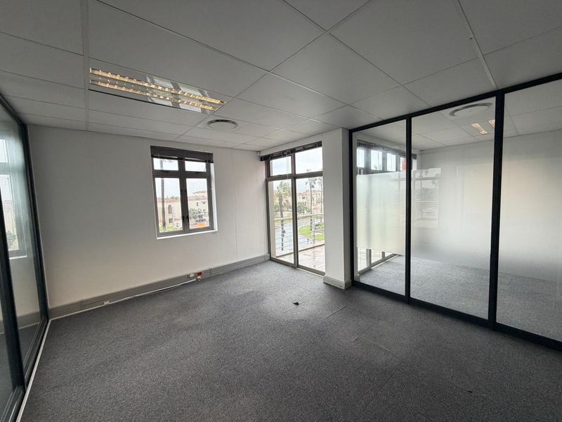 A bright, vacant commercial office space featuring glass partitioning, suspended ceiling tiles, and large windows providing natural light and exterior views.