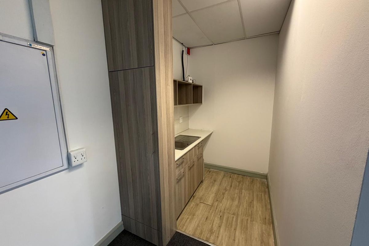 A compact kitchenette area featuring wood-look cabinetry, a built-in sink, and overhead shelving, suitable for a small office or commercial utility space.