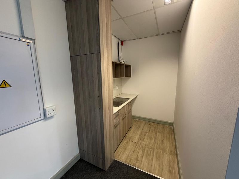 A compact kitchenette area featuring wood-look cabinetry, a built-in sink, and overhead shelving, suitable for a small office or commercial utility space.
