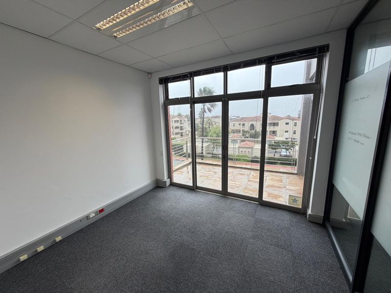 A bright, empty office space featuring carpeted flooring, a suspended ceiling with recessed lighting, and large glass doors opening onto a private balcony.