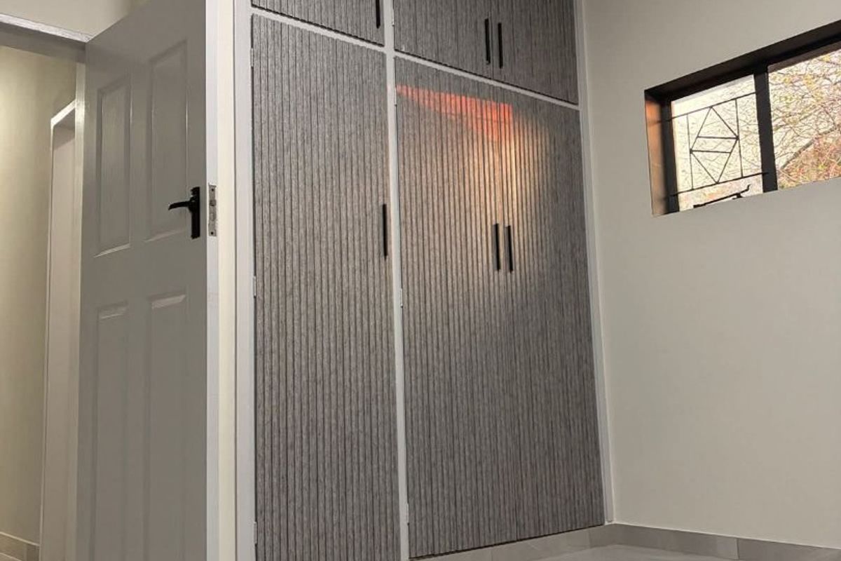 A clean, modern bedroom featuring floor-to-ceiling built-in cupboards with textured grey doors, polished tiled flooring, and a secure window.
