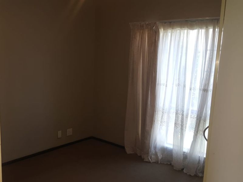 An empty bedroom featuring neutral walls, carpeted flooring, and a window with sheer curtains, offering a blank canvas for personalization.