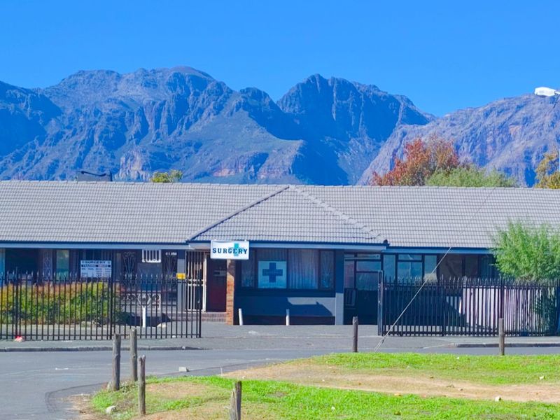 Front view of a commercial surgery building featuring a tiled roof, secure perimeter fencing, and a paved driveway with mountain views in the background.