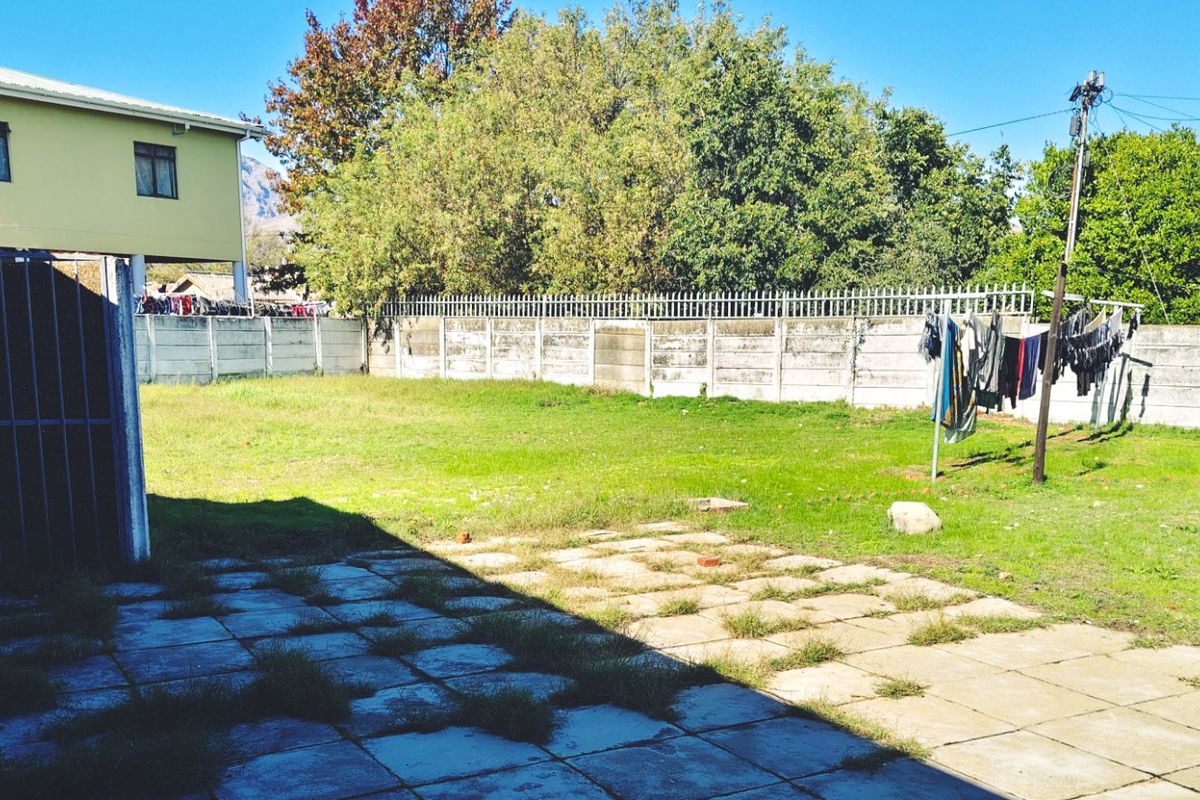 A rear exterior view of the property featuring a paved patio area, a grass lawn, and a precast concrete boundary wall with security spikes.