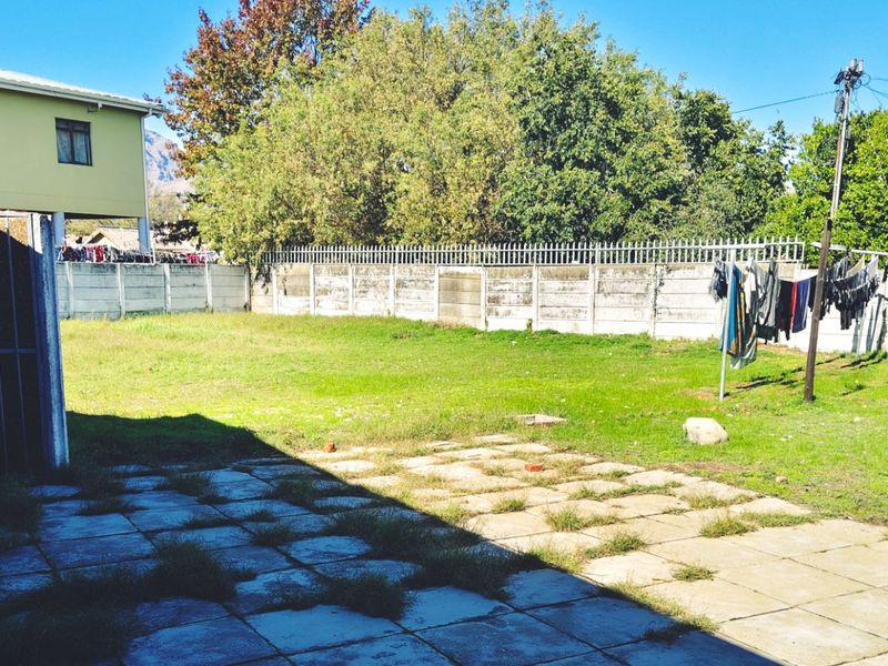 A rear exterior view of the property featuring a paved patio area, a grass lawn, and a precast concrete boundary wall with security spikes.