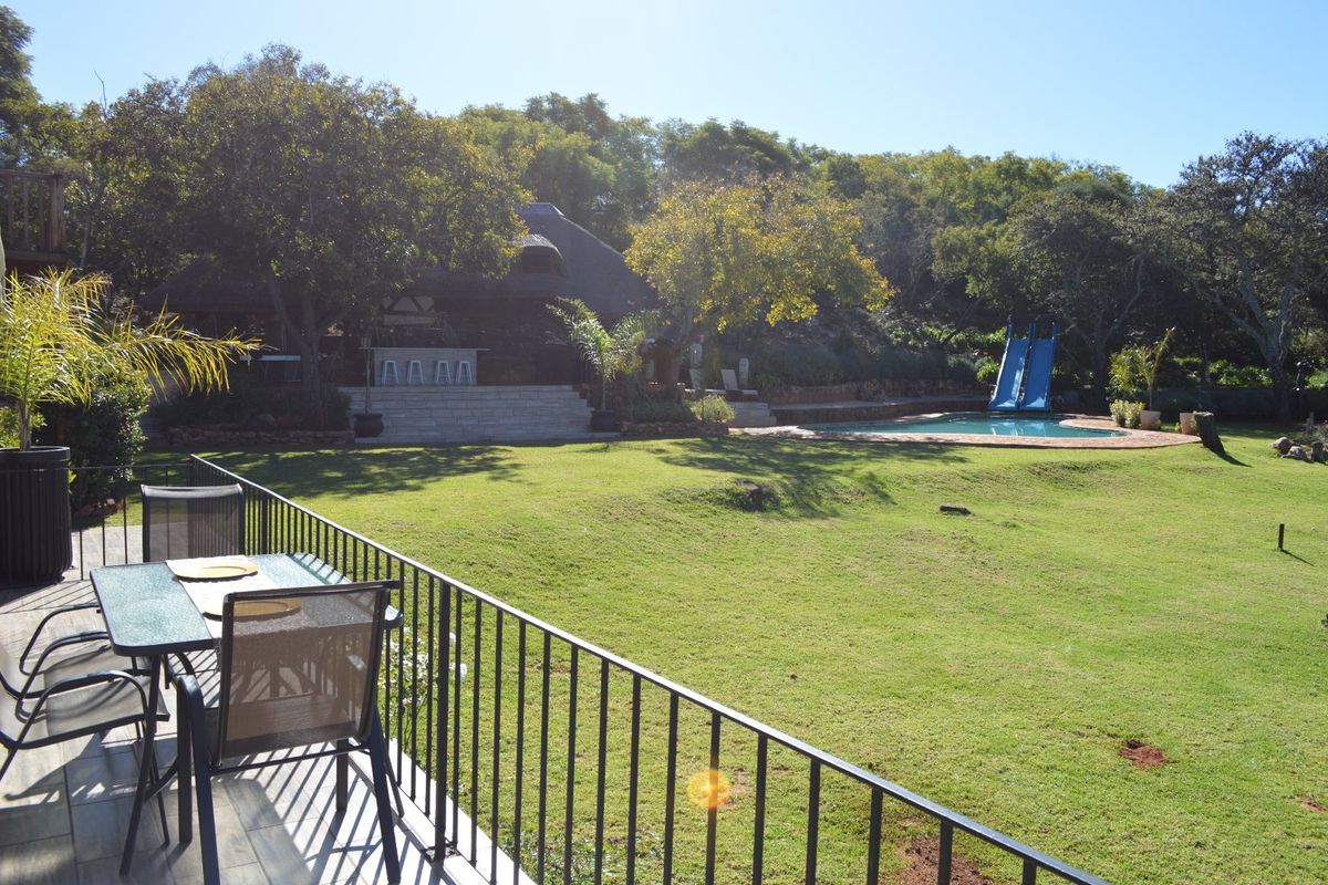 Spacious rear garden featuring a swimming pool with a slide, stone steps leading to a thatched entertainment area, and a tiled balcony with metal railings.