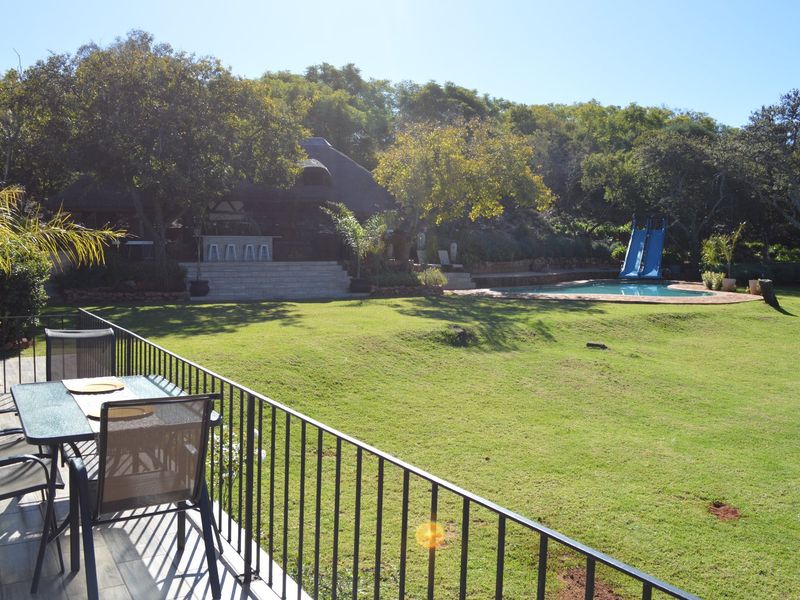 Spacious rear garden featuring a swimming pool with a slide, stone steps leading to a thatched entertainment area, and a tiled balcony with metal railings.