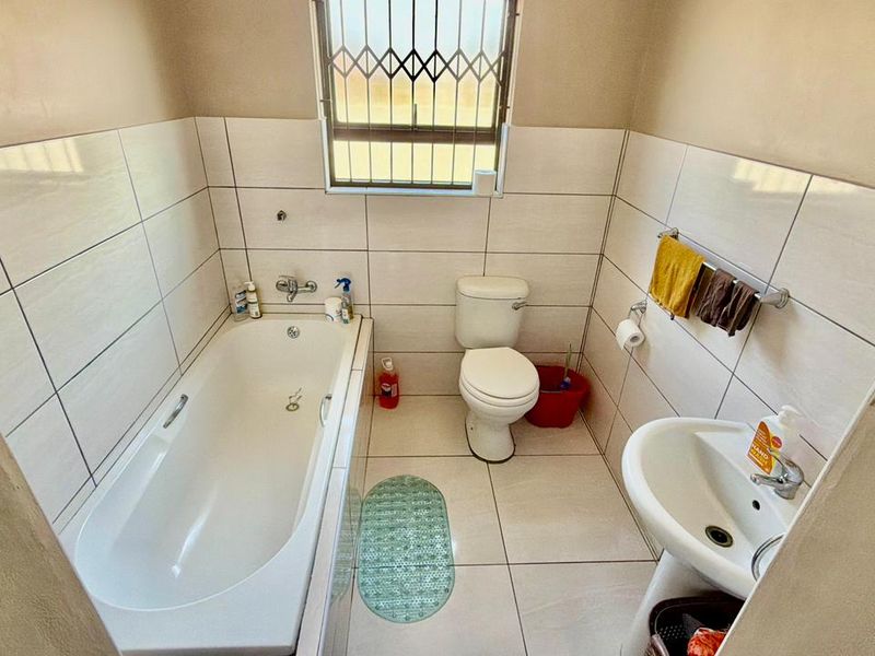A functional bathroom featuring a bathtub, pedestal basin, and toilet, finished with neutral wall and floor tiling and a security-barred window.