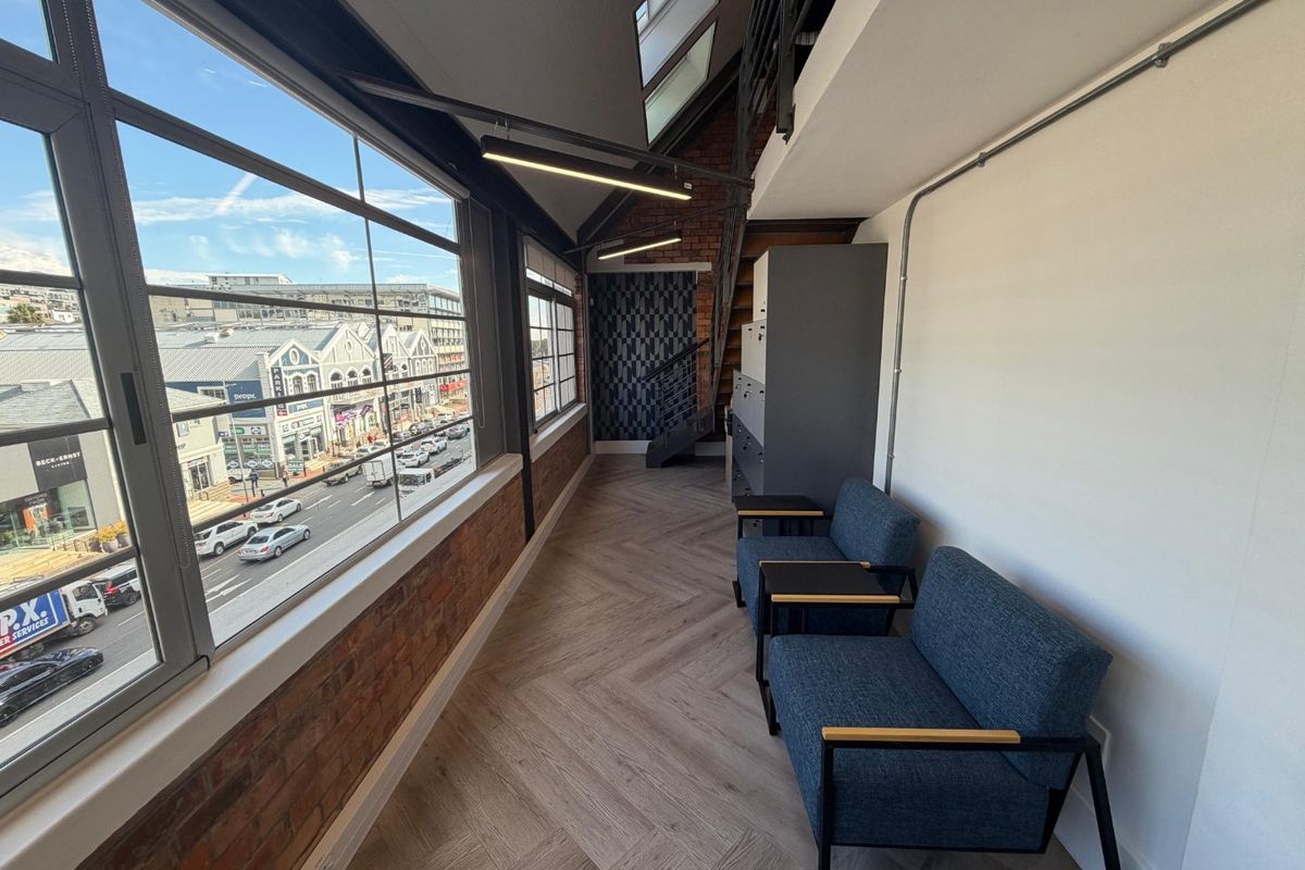 Modern industrial-style office space featuring exposed brickwork, herringbone flooring, skylights, and large windows offering an urban street view.