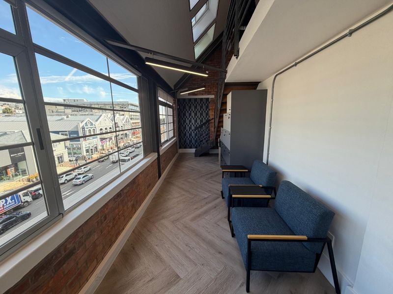 Modern industrial-style office space featuring exposed brickwork, herringbone flooring, skylights, and large windows offering an urban street view.