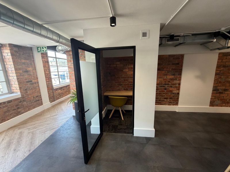 Modern commercial office space featuring exposed brick walls, industrial-style exposed ducting, herringbone flooring, and a private built-in work pod.