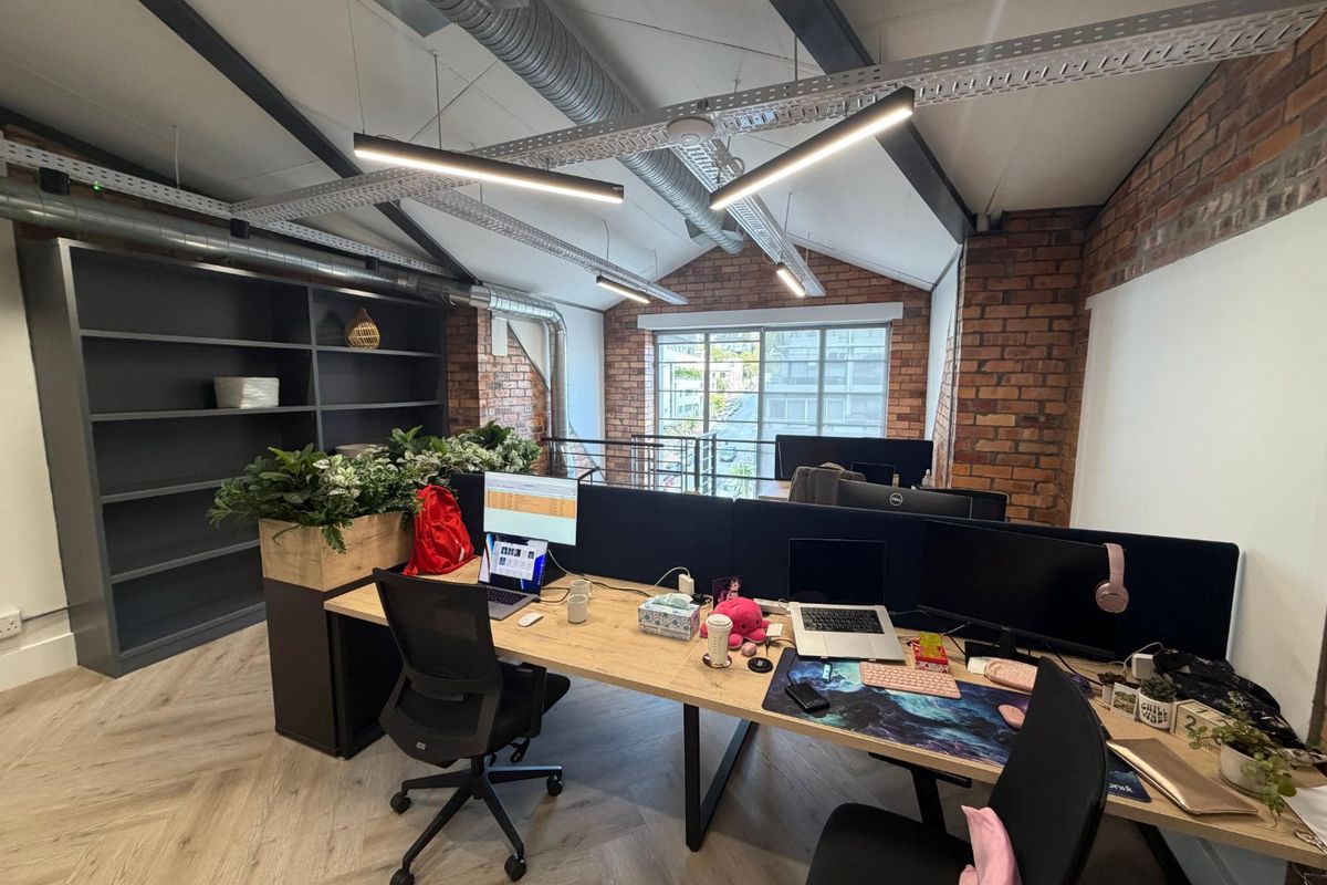 Modern industrial-style office space featuring exposed brick walls, vaulted ceilings with exposed ductwork, herringbone flooring, and built-in shelving.
