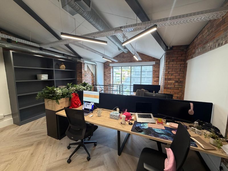Modern industrial-style office space featuring exposed brick walls, vaulted ceilings with exposed ductwork, herringbone flooring, and built-in shelving.