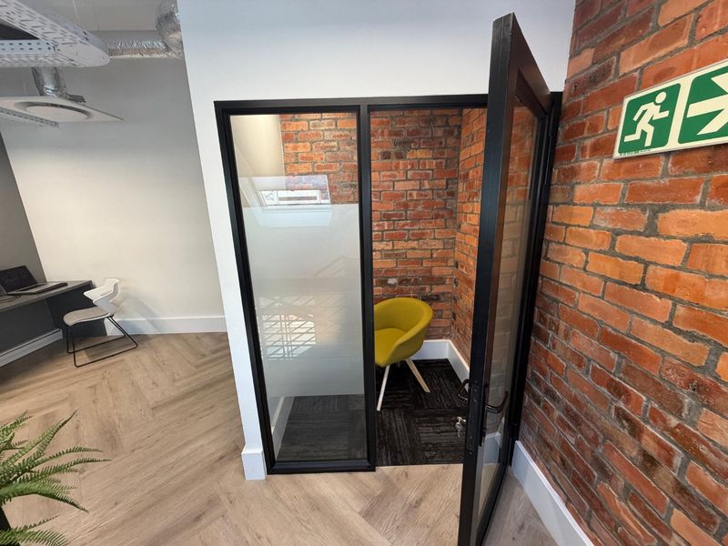 A modern office pod featuring exposed brick walls, glass partitioning, and contemporary herringbone flooring within a professional commercial workspace.