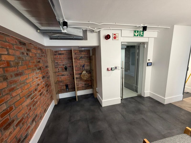 Modern commercial hallway featuring exposed brickwork, industrial-style exposed ducting, tiled flooring, and a secure glass entrance door with integrated fire safety systems.