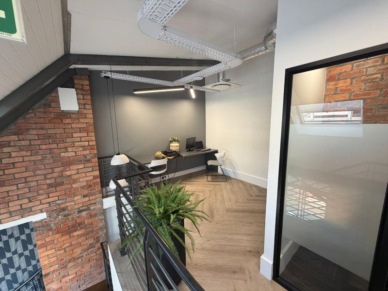 Modern commercial office space featuring herringbone timber flooring, exposed brickwork, industrial-style cable trays, and a built-in desk area with glass partitioning.