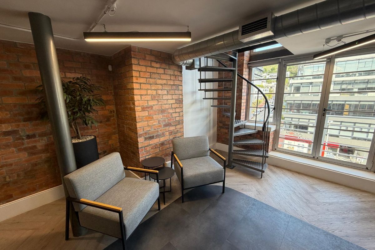 Modern office space featuring exposed brick walls, industrial-style spiral staircase, exposed ducting, and large windows providing ample natural light.