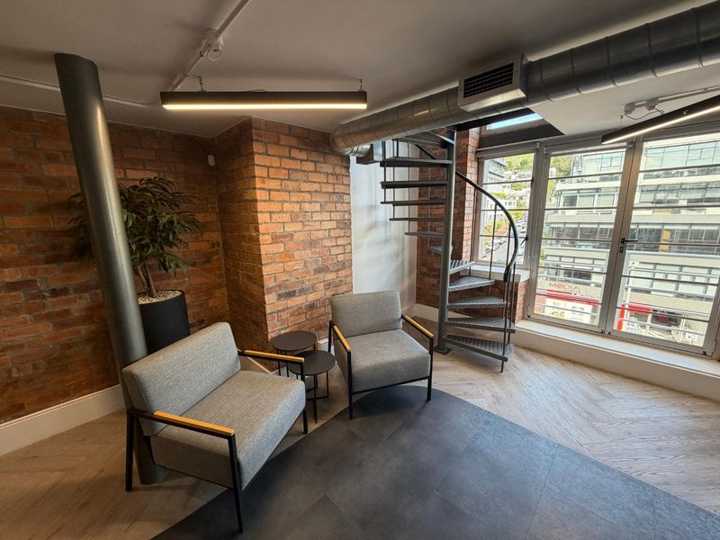 Modern office space featuring exposed brick walls, industrial-style spiral staircase, exposed ducting, and large windows providing ample natural light.