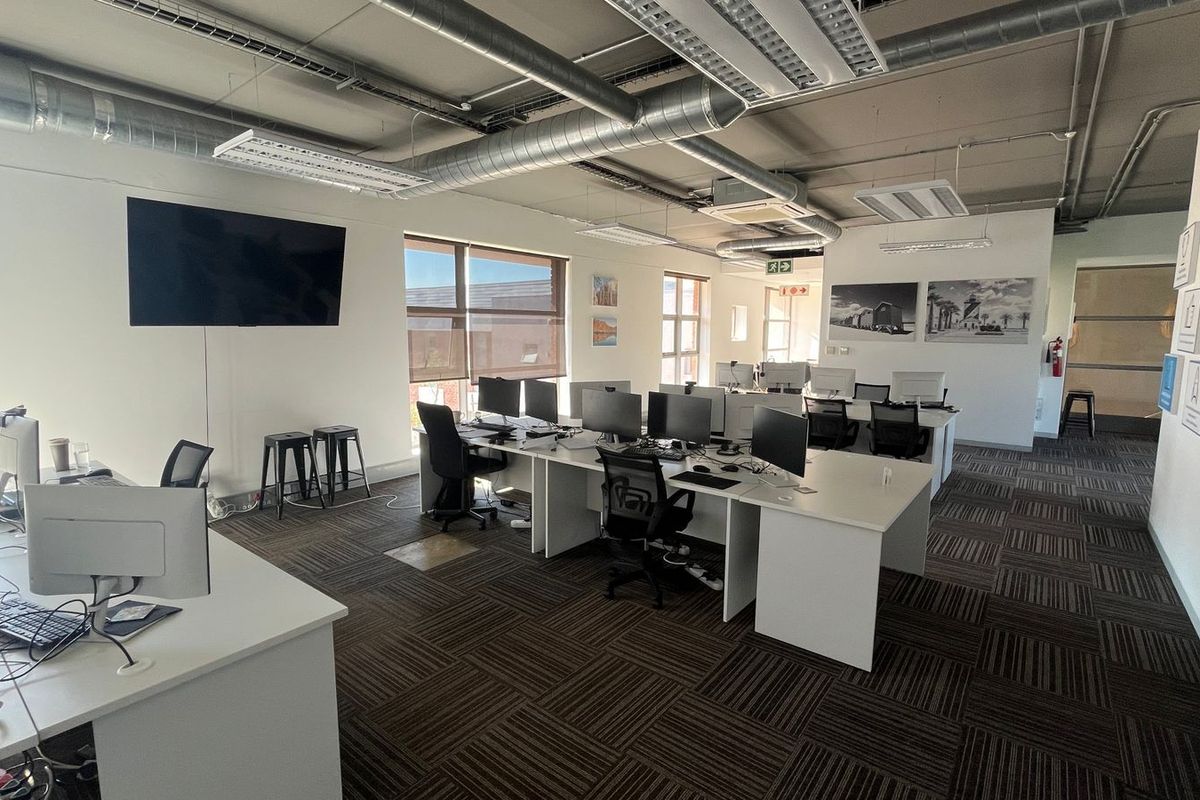 Open-plan commercial office space featuring exposed industrial ducting, carpet tile flooring, and large windows providing ample natural light.