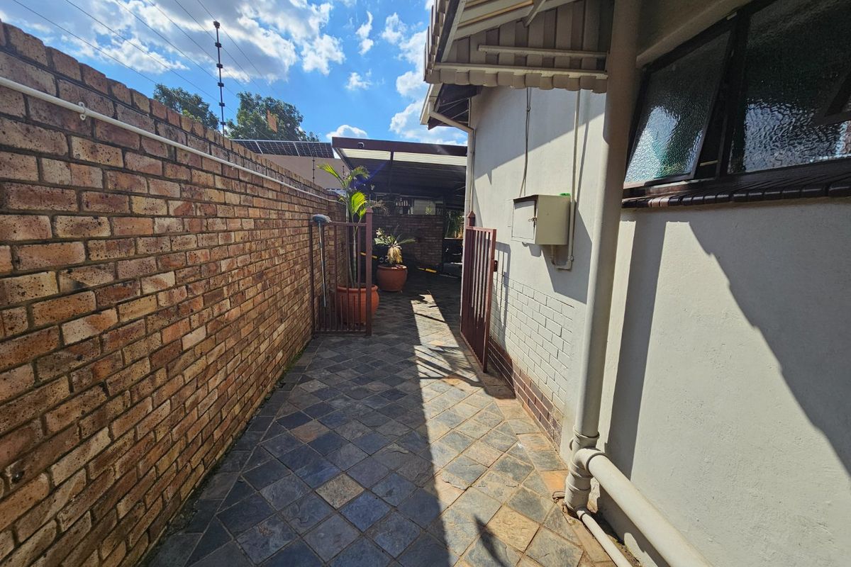 A side walkway featuring a brick boundary wall, paved flooring, and a metal gate leading to the rear outdoor area.